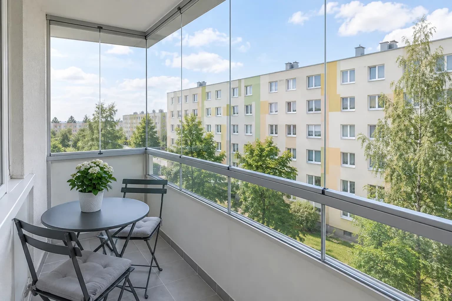 Przeszklony balkon w bloku: szklane tafle w cienkich aluminiowych ramach, stolik i roślina w środku