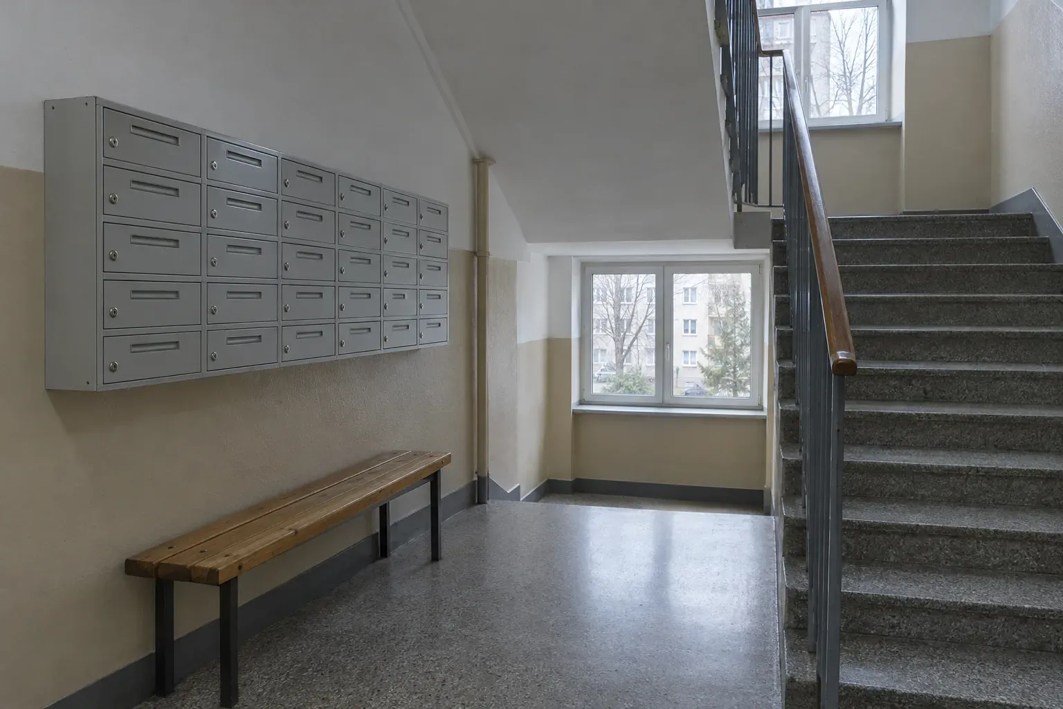 Wejście do klatki schodowej bloku mieszkalnego z rzędem skrzynek pocztowych i drewnianą ławką